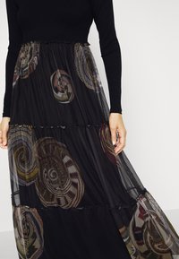 Abito maxi con un leggero strato esterno nero con motivi ondulati di vari colori. Corpino aderente con maniche lunghe e vita arricciata.