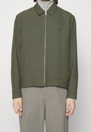 Modèle portant une veste zippée vert olive sur un pull à col roulé gris clair et un pantalon gris ample, debout devant un fond uni.