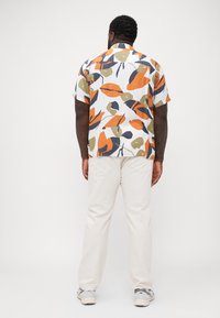 Camisa de botones de manga corta en blanco, con un patrón abstracto de hojas en naranja, verde y azul marino. Se lleva con pantalones color crema.