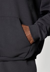 Sudadera gris oscuro con una textura suave, que cuenta con un bolsillo tipo canguro y puños acanalados. Detalles de cuero marrón visibles en la mano.