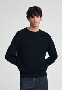 Zwarte, geribbelde gebreide trui met een ronde halslijn en lange mouwen. Heeft een aansluitende zoom en subtiele zijsplitten, gecombineerd met donkere jeans.