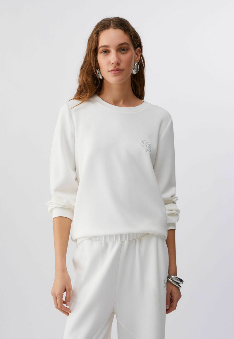 Sweatshirt blanc en tissu doux, avec un col rond, une coupe ample, et un détail de logo brodé. Associé à un pantalon blanc assorti.