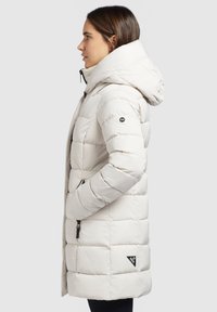 Leichte, cremefarbene Steppjacke mit Kapuze, genoppter Textur, schwarzem Logo auf dem Ärmel und mit Reißverschluss versehenen Seitentaschen.