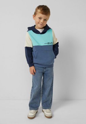 Chłopiec stoi z dłońmi w kieszeniach, ubrany w niebiesko-kremową bluzę, luźne niebieskie dżinsy i białe trampki, na tle gładkiego szarego tła.