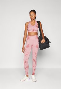 Rosa tie-dye sports-bh og tights-set, med en glatt tekstur. Modellen holder en svart treningsbag. Hvite joggesko med lilla detaljer.