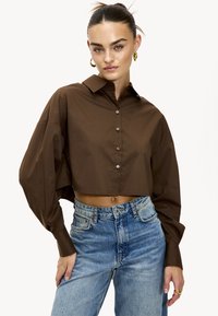 Chemise courte marron à boutons avec un col et des manches longues et larges. Associée à un jean taille haute en délavé clair.