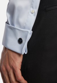 Puño de camisa de vestir azul claro con cierre de botón y acento circular en navy, con textura de tela suave y corte ajustado.