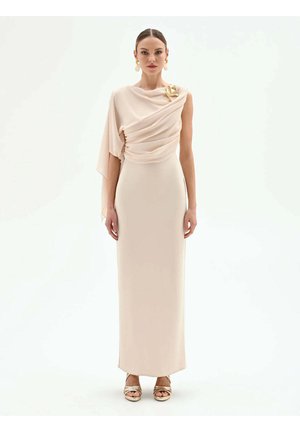 Donna che indossa un lungo abito beige asimmetrico con tessuto drappeggiato su una spalla, spilla dorata, orecchini dorati e sandali aperti dorati.