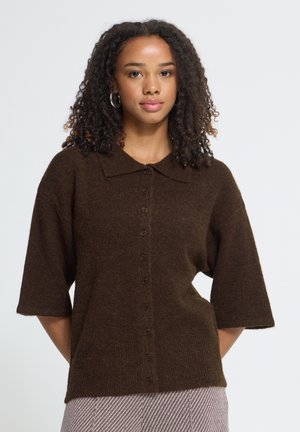 Femme aux cheveux bouclés portant un pull boutonné à manches courtes marron foncé, debout devant un fond clair et uni, regardant devant elle.