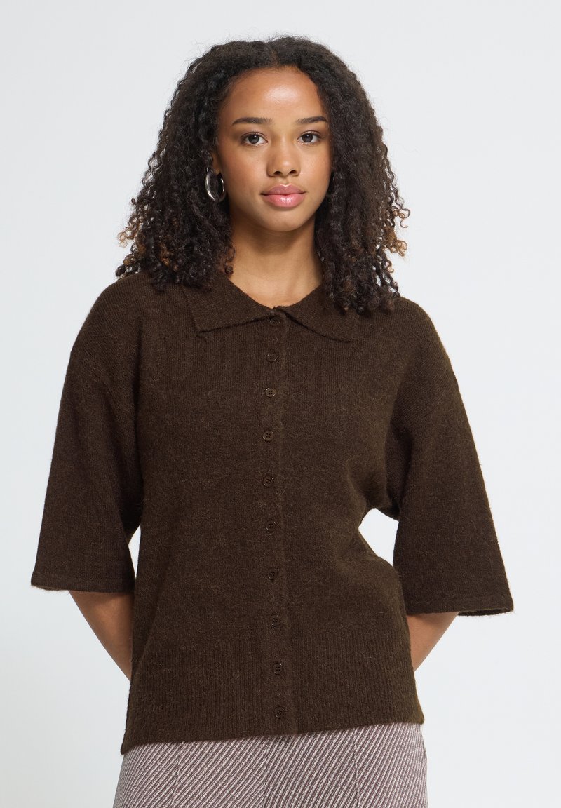 Femme aux cheveux bouclés portant un pull boutonné à manches courtes marron foncé, debout devant un fond clair et uni, regardant devant elle.