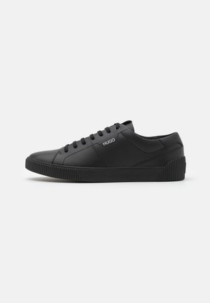 Zwarte lage sneaker met zwarte veters, gestructureerde zool en "HUGO" logo aan de zijkant, getoond in zijaanzicht op een effen achtergrond.