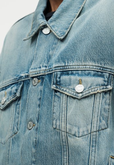 Κοντινό πλάνο ανοιχτό μπλε denim μπουφάν που δείχνει τσέπες στο στήθος με κουμπωτές φλος και ασημένια μεταλλικά κουμπιά με λεπτομέρειες ραφής.