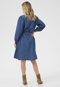 Sininen farkkumekko, jossa on pitkät puff-hihat, kiristetty vyötärö ja polvimittainen helma. Yksityiskohtana napilliset hihansuut ja ompeluaksentit.