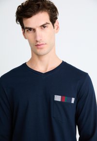 Maglietta Navy a maniche lunghe con scollo a V realizzata in tessuto morbido, caratterizzata da una tasca con motivo a righe in grigio, rosso, verde e navy.