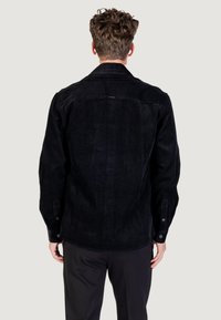 Giacca in corduroy nera con colletto classico, maniche lunghe e polsini con bottoni; presenta una texture a righe verticali e una vestibilità rilassata.