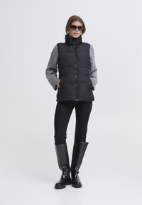 Chaleco acolchado negro con cuello alto sobre manga larga gris, combinado con leggings negros y botas negras hasta la rodilla. Gafas de sol incluidas.