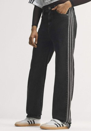 Person iført sorte jeans med grå sidelister, en gennemsigtig sort langærmet top under en hvid skjorte og hvide sneakers med sorte striber.