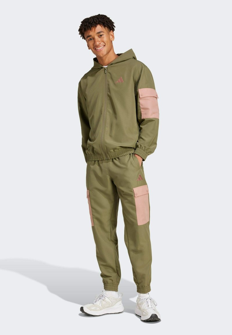 Olive green tracksuit with a zip-up hoodie and elastic cuffs, featuring pink accents on the sleeves and pants. Made from lightweight material.