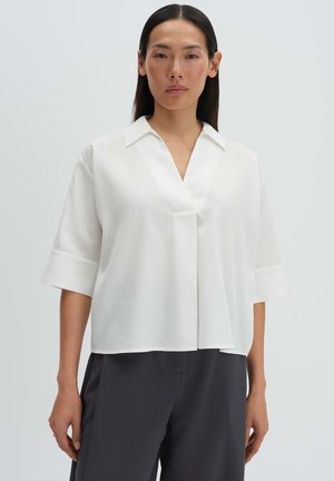 Frau trägt eine locker sitzende weiße Kurzarmbluse mit V-Ausschnitt und dunkel weite Hosen, steht vor einem schlichten Hintergrund.