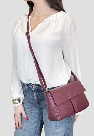 Frau trägt weiße Bluse und blaue Jeans, dazu eine strukturierte crossbody Tasche aus bordeauxrotem Leder mit verstellbarem Riemen.