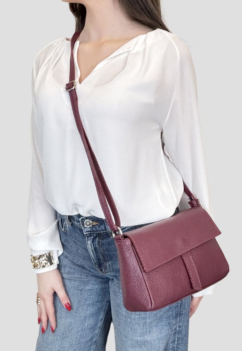 Donna con camicetta bianca e jeans blu, che porta una borsa a tracolla in pelle bordeaux con trama e tracolla regolabile.