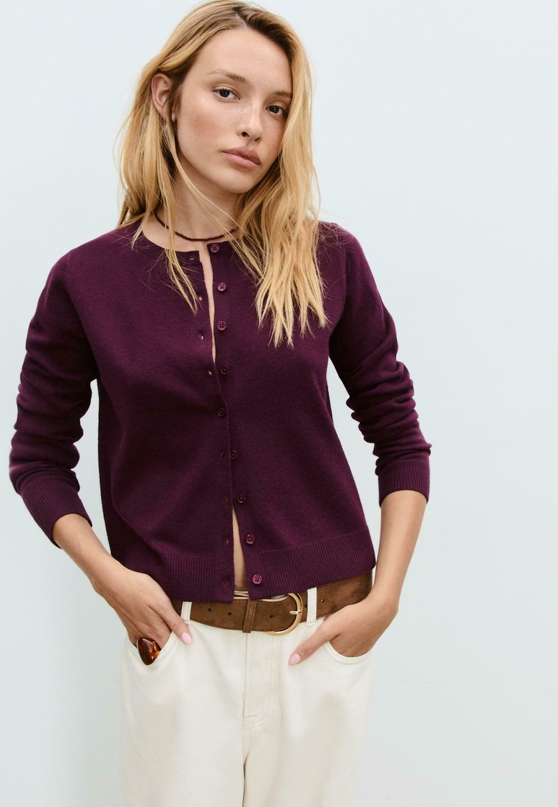 Cardigan bourgogne à manches longues, encolure ronde et boutons sur le devant. Porté par-dessus un pantalon crème, avec une ceinture marron pour le contraste.