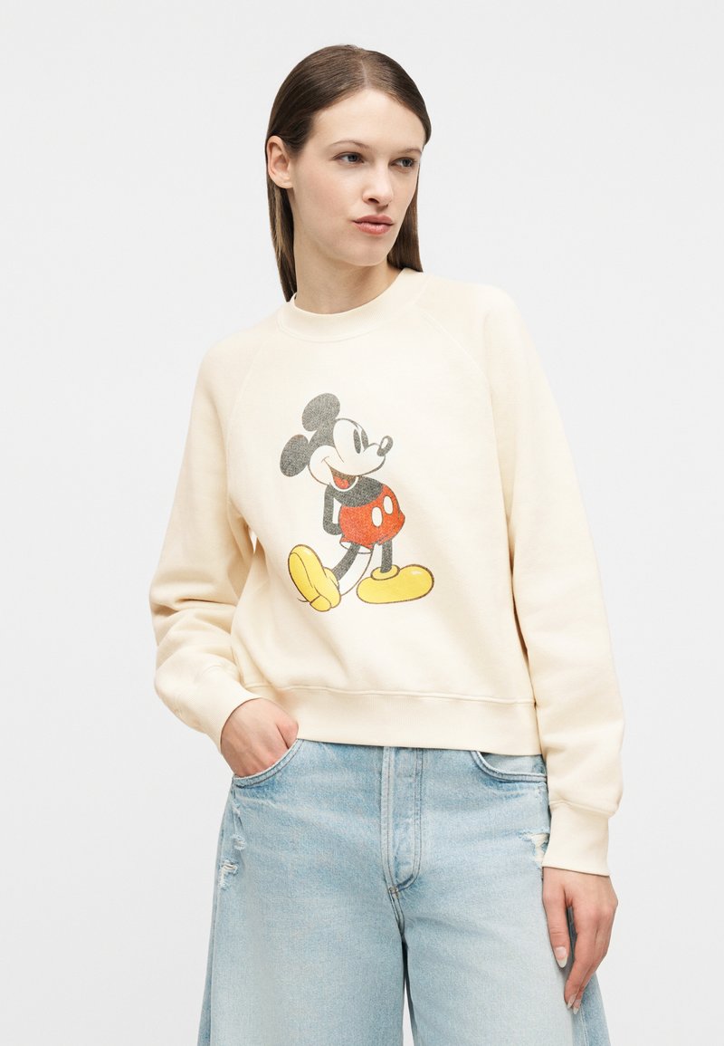 Femme portant un sweat-shirt crème avec un graphisme vintage de Mickey Mouse et un jean bleu clair déchiré, debout devant un fond uni.