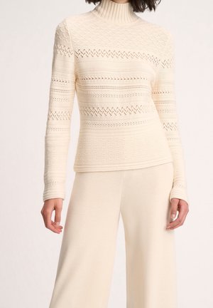 Woman wearing a cream-colored knit turtleneck sweater with patterned holes and matching wide-leg pants against a neutral background.