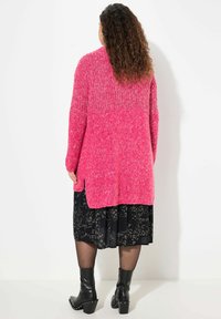 Cardigan rosa lavorato a maglia con una parte superiore testurizzata, retro più lungo, spacco laterale, indossato su una gonna nera a fantasia e abbinato a stivali neri con tacco.