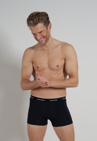 Schwarze Boxershorts aus weichem Stoff, mit einem Bund, der mit "ceceba"-Branding versehen ist. Das Modell steht mit gefalteten Händen und lächelt.