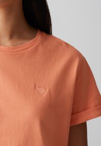 T-shirt in cotone arancione chiaro a maniche corte e collo rotondo, con un piccolo cuore ricamato sul lato sinistro del petto. Texture liscia.