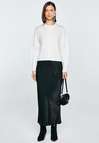 Bílý, zapínací vlněný kardigan s dlouhými rukávy, ve spojení s černou saténovou midi sukni, černými kotníkovými botami a malou černou kabelkou.