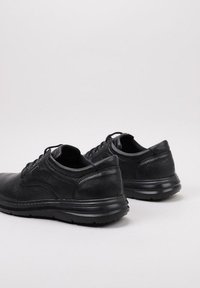 Par de zapatos de cuero negro con cordones, superficie texturizada y suelas de goma gruesas sobre un fondo claro.