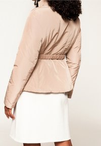 Ljust beige puffer-jacka med bälte i midjan, kviltad textur och en något kortad design. Slätt tyg med hög krage.