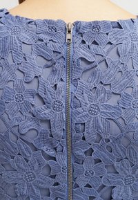 El vestido de encaje floral azul presenta en la parte trasera una cremallera con dientes de metal. Un intrincado bordado muestra un patrón de flores sobre un fondo de tela transparente.