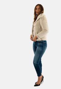 Chaqueta de pelo sintético en color crema con capucha, superficie texturizada y cierre frontal de botones, combinada con pantalones vaqueros azules y tacones altos negros.