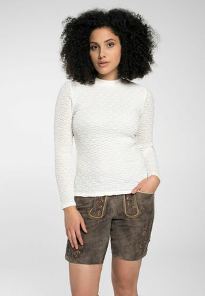 Weißes Langarmoberteil aus strukturiertem Stoff, kombiniert mit braunen Wildleder-Shorts, die mit floralen Stickereien und Taschen versehen sind.