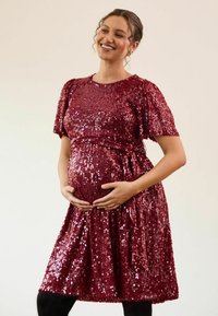 Punane sädelev raseduskleit, millel on puhvärviga varrukad ja taljepael, flared seeliku ja ümar neckline, kantud mustade sukkpükstega.