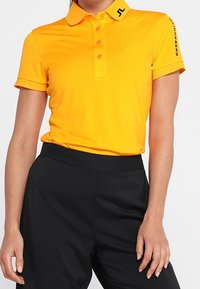 Kortärmad, collared polo skjorta i starkt gult, med fem knappar, svarta logotyper och ett mjukt, elastiskt tyg. Matchad med svarta byxor.