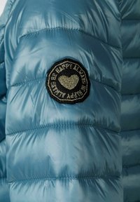 Ljusblå puffjacka med diagonala sömmar och en rund svart patch med en broderad hjärta och text.