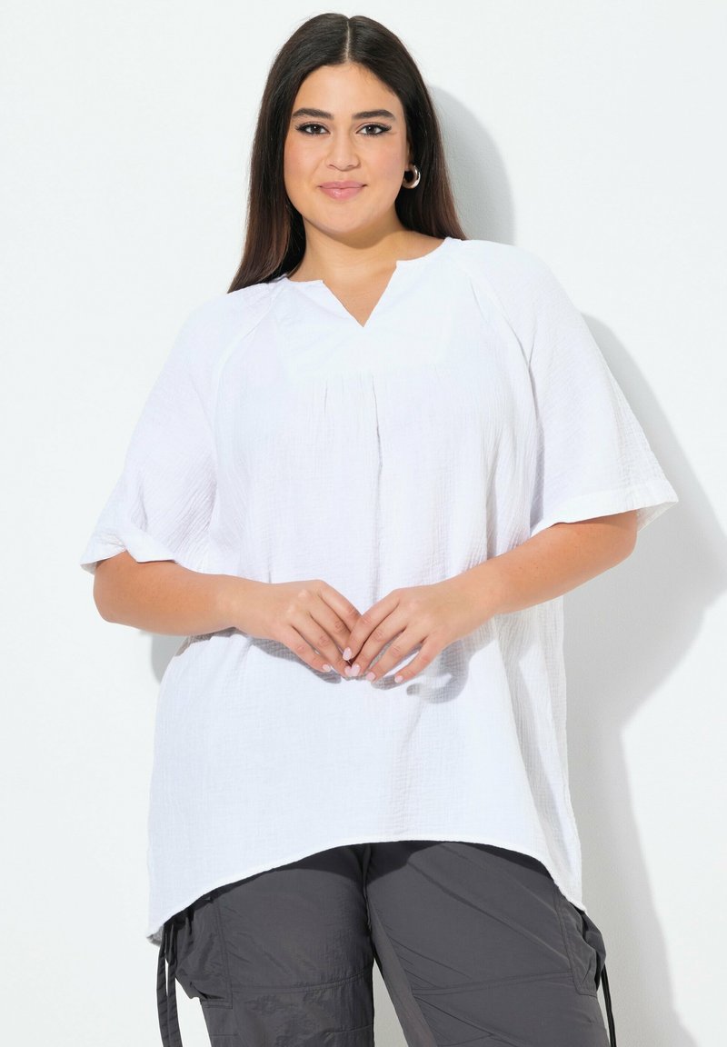 Top bianco a maniche corte in un tessuto testurizzato con scollo a V e dettaglio di piega. L'orlo è più lungo sul retro. Abbinato a pantaloni cargo grigi.