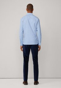 Man die met zijn rug naar de camera staat, gekleed in een lichtblauw geruit overhemd met lange mouwen, donkere marineblauwe broek en donkerbruine schoenen, tegen een neutrale achtergrond.