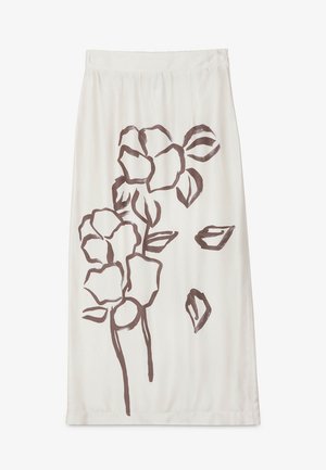 Vrouw draagt een beige blazer, bruin strapless topje, lange crèmekleurige rok met grote donkere bloemenprint en beige sandalen met hak, staand tegen een lichte achtergrond.