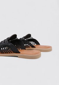 Sandalias negras trenzadas con un diseño de punta abierta, que cuentan con una plantilla en color marrón claro y una suela de color claro. Textura suave y detalle intrincado en las correas.