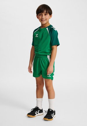 Kind in groene sportkleding met witte accenten en zwarte binnensportschoenen, staand en glimlachend tegen een effen achtergrond.