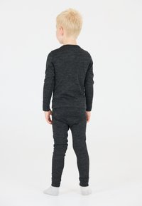 Blond kind dat blootsvoets met de rug naar de kijker staat, gekleed in een donkergrijze langemouwen top en bijpassende broek tegen een witte achtergrond.