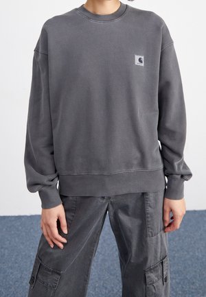 Personne portant un sweat-shirt Carhartt gris charbon délavé et un pantalon cargo assorti, debout, les mains détendues le long du corps.