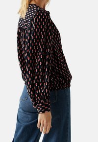 Blusa nera con motivo a diamante rosso e bianco, maniche lunghe e orlo elasticizzato. Abbinata a jeans in denim blu chiaro.