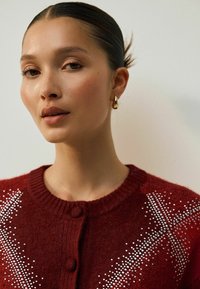 Červený pletený cardigan s kulatým výstřihem, dvěma knoflíky a stříbrnými ozdobami vytvářejícími tečkovaný vzor na přední straně. Hladká textura.