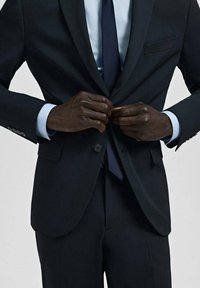 Donker marineblauw pak met een omgeslagen kraag, nauwsluitende broek en een jasje met knopen. Gecombineerd met een lichtblauw overhemd en bijpassende stropdas.